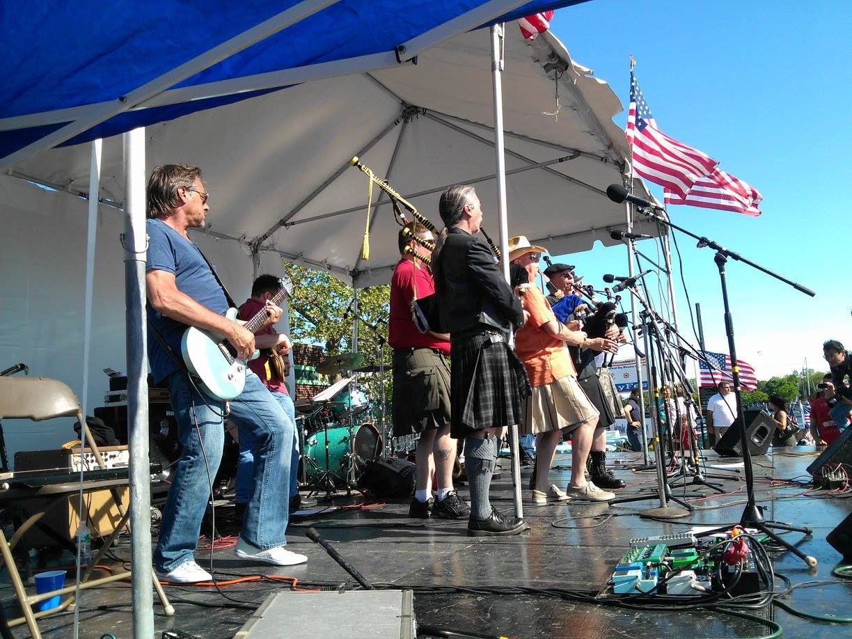 Band performing on an outdoor stage under a tent, with guitars, microphones, and an American flag nearby.