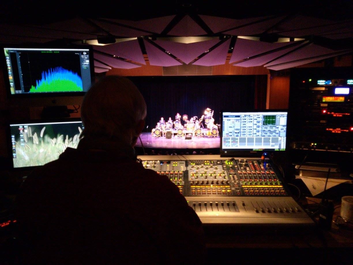 Sound engineer at a mixing console watches a lit stage with performers and audio monitors in a dark venue