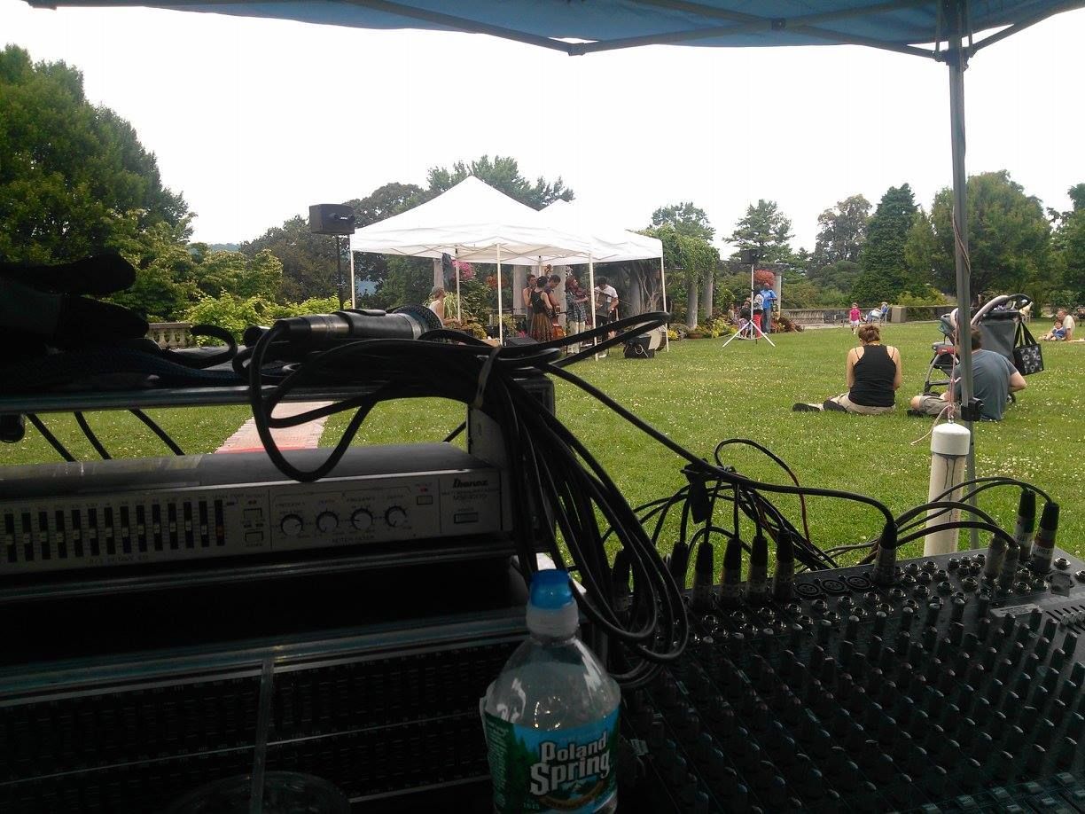 Outdoor concert under a canopy, with lawn seating, tents, and people gathered in a park.