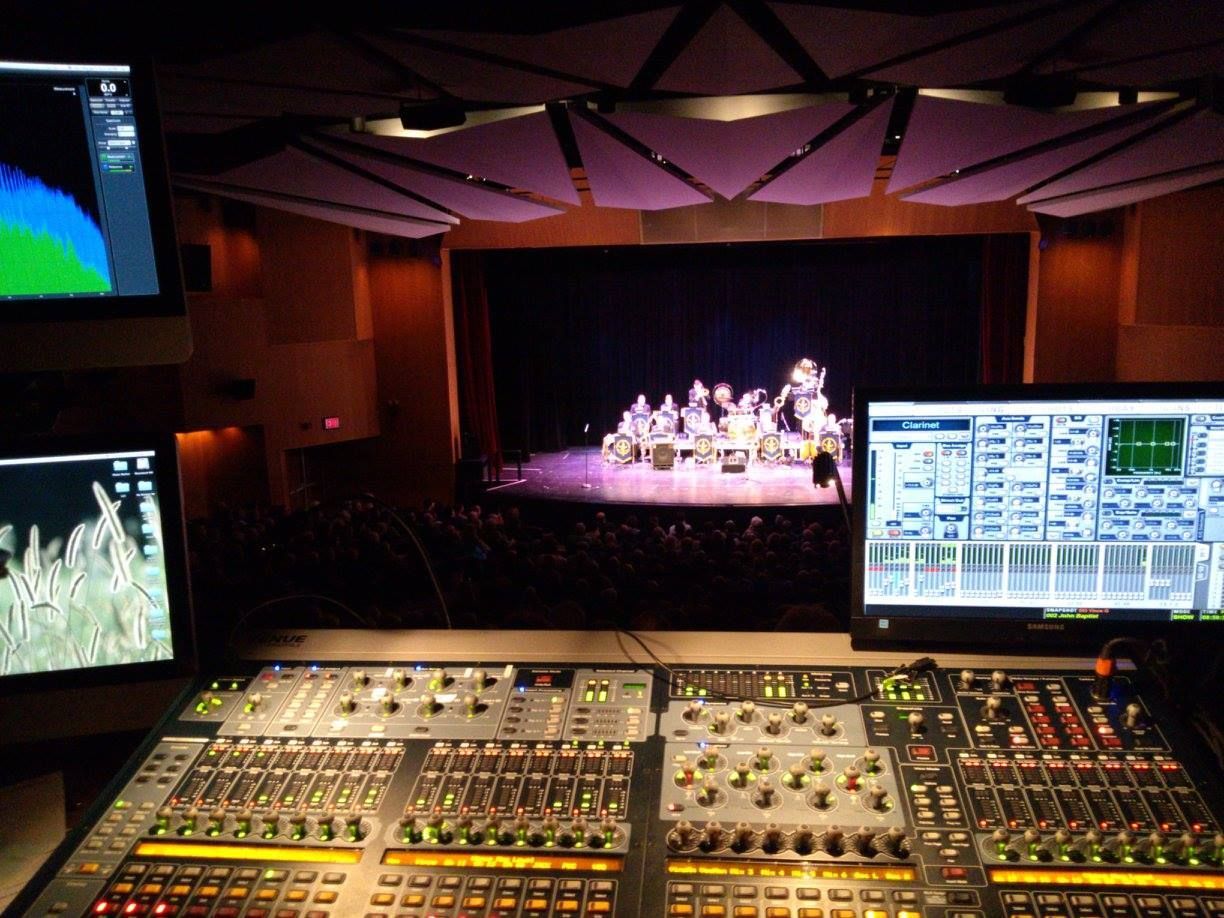 Sound mixing console in a control room, overlooking a lit stage rehearsal with performers.