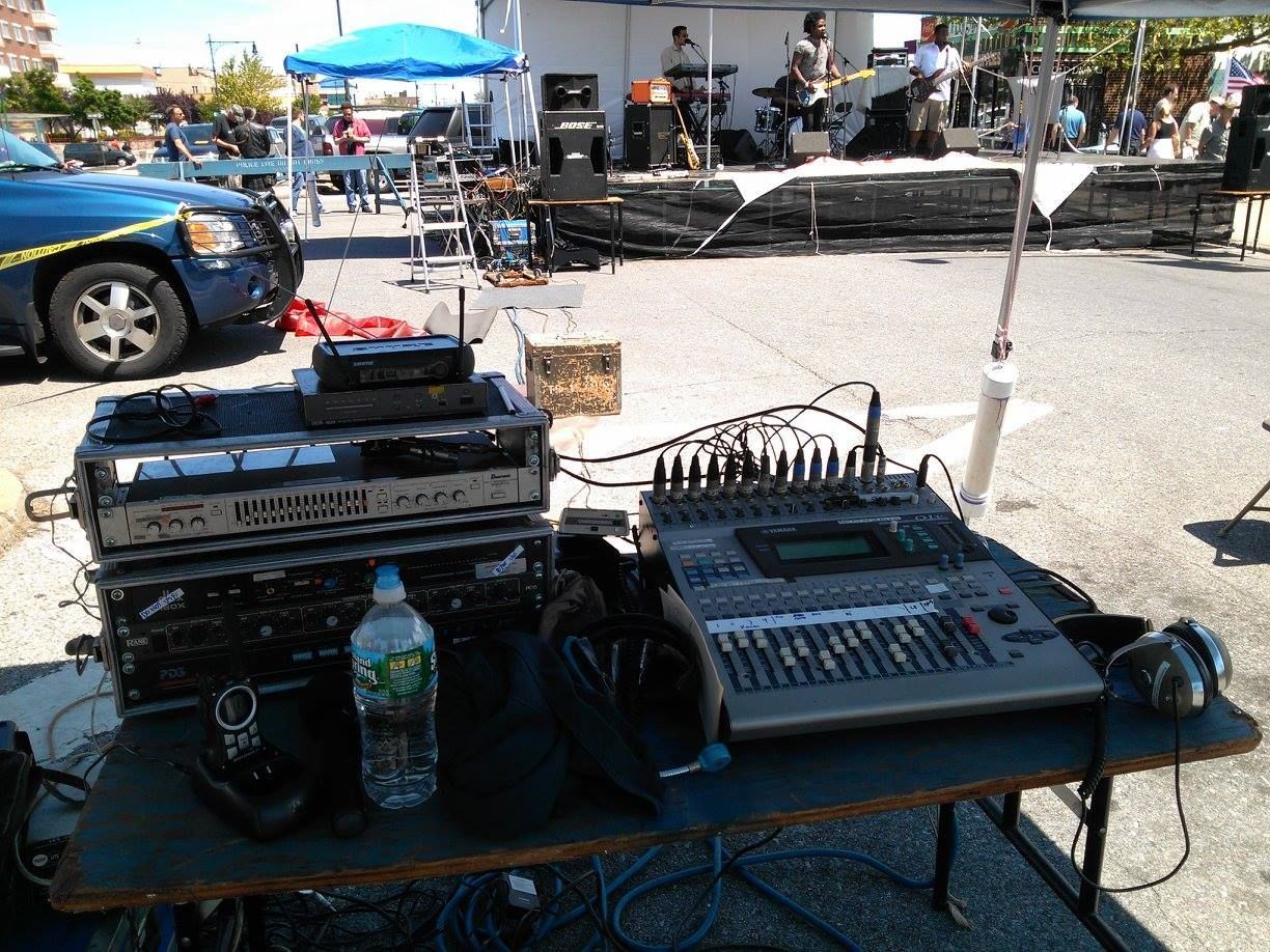 Audio mixing setup on a table at an outdoor event, with speakers, cables, and a blue car nearby