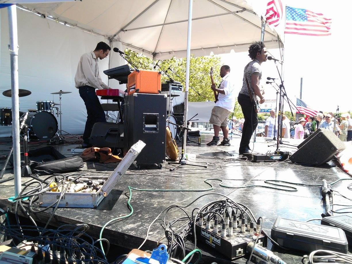 Band performing on an outdoor stage under a white canopy, with microphones, amps, and cables on the floor.