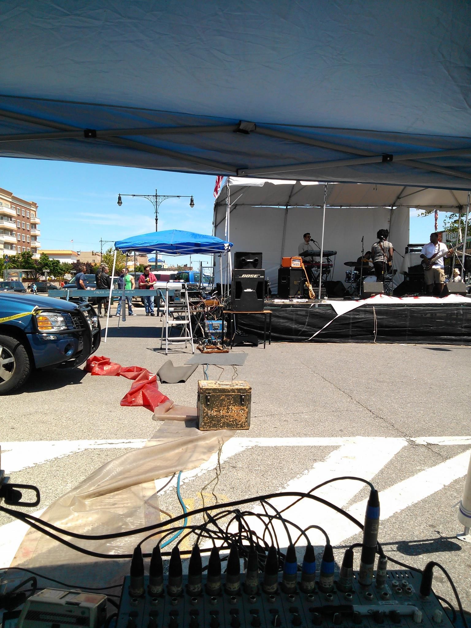 Outdoor live music stage under a blue canopy, with audio equipment in the foreground and a parked car at left.