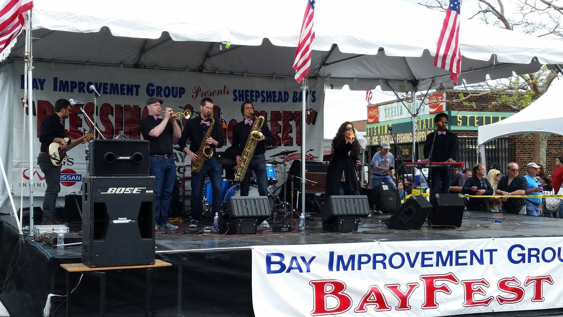 Band performing on an outdoor stage at Bay Fest under white tents, with Bay Improvement Group banner in front.