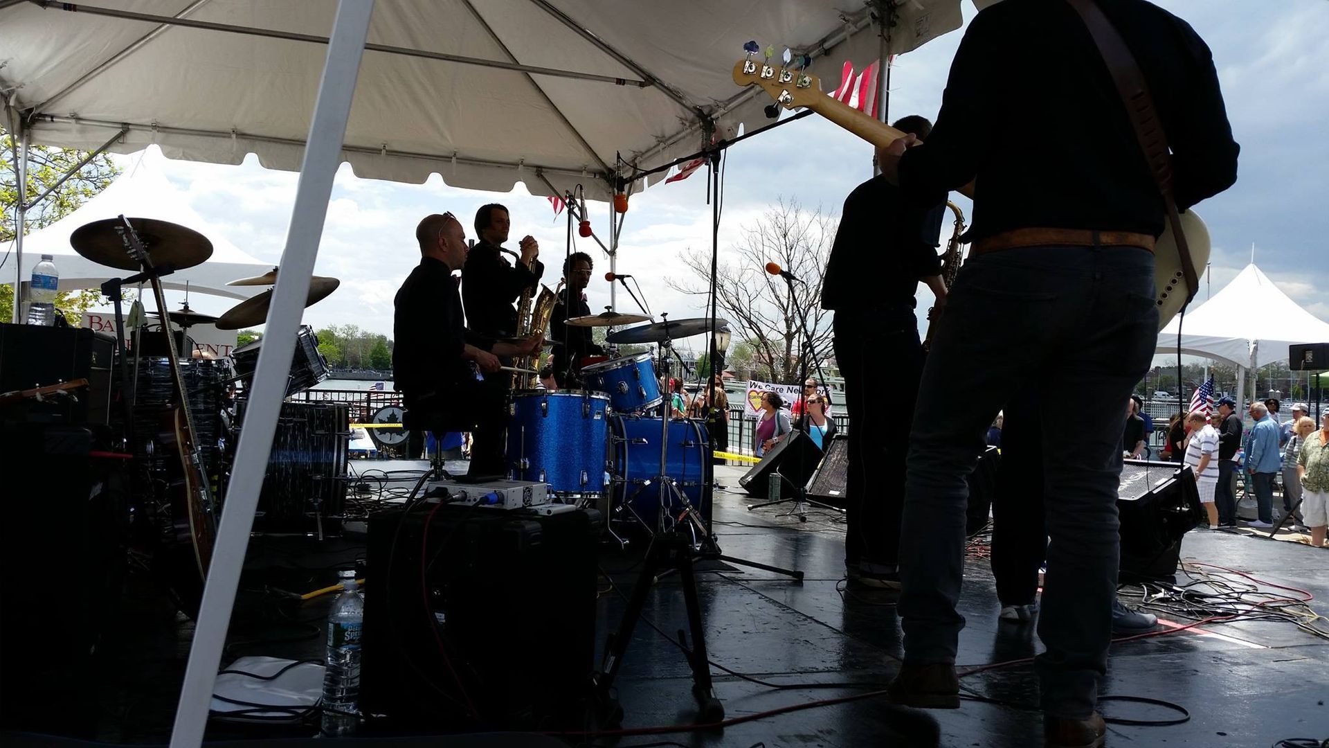 Band performing on an outdoor stage under a white tent, with drums and audience visible.