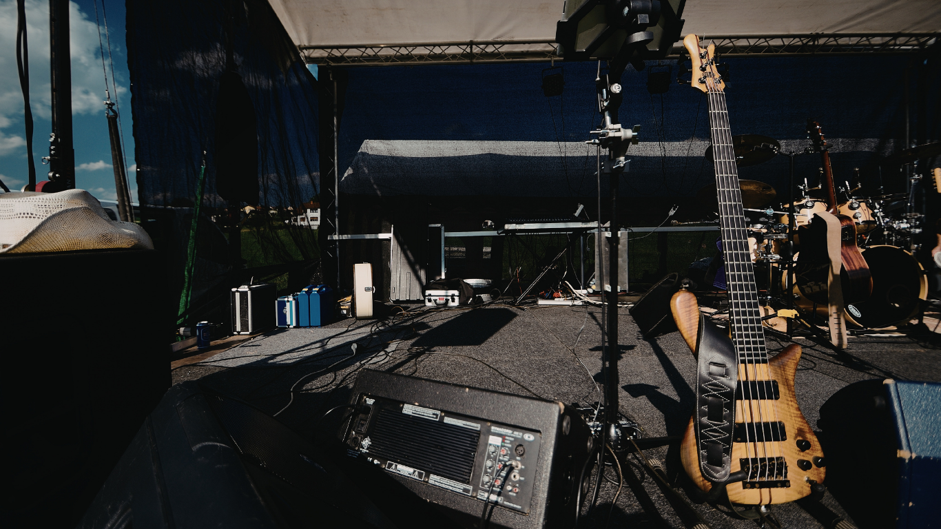 Outdoor stage setup with microphone stand, speakers, and guitars in front of a covered performance area