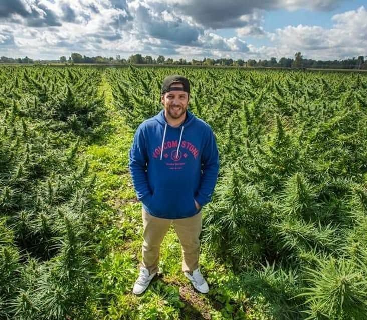 Man standing in a lush green field of cannabis plants, wearing a blue hoodie and smiling