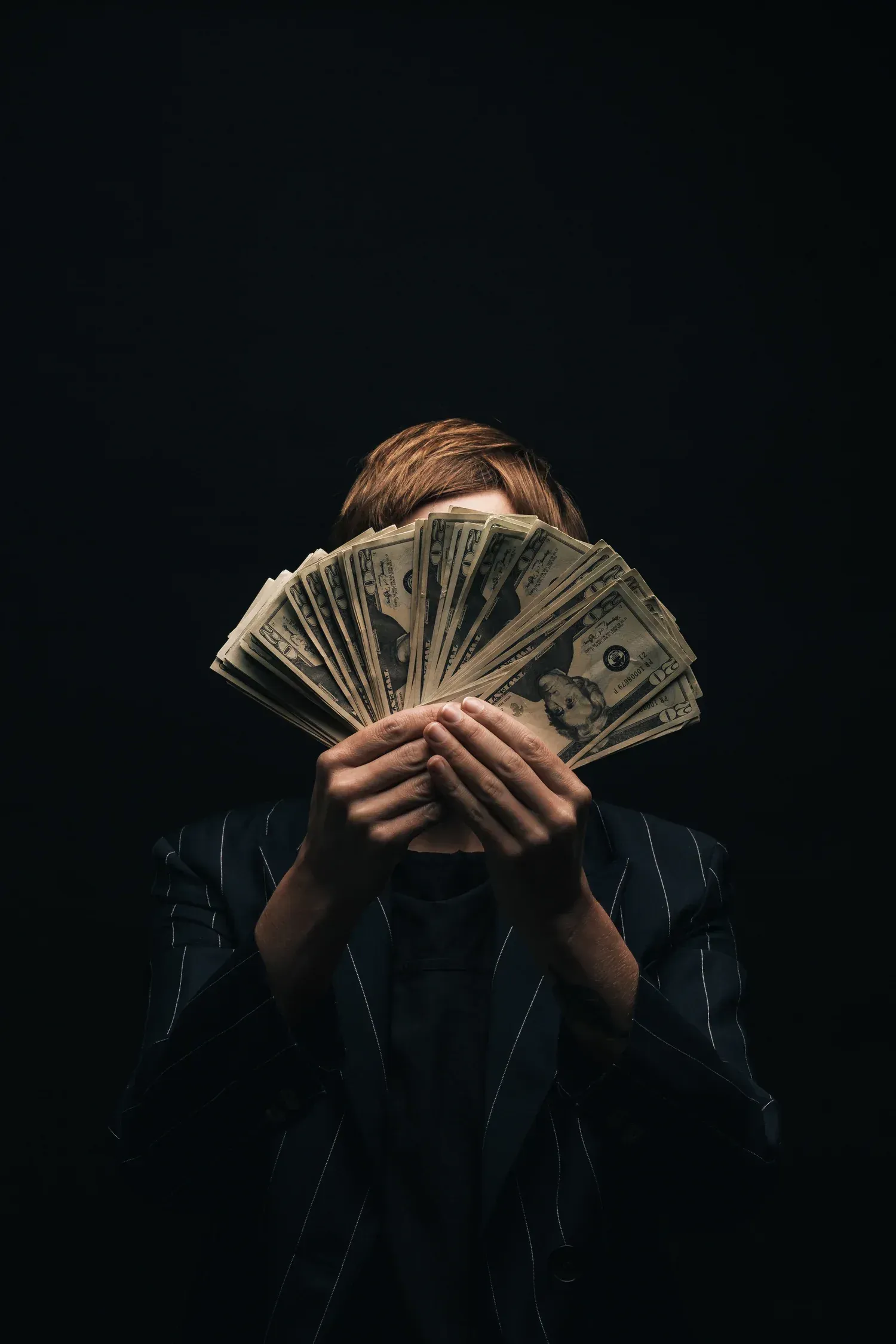 Person holding a fan of cash in front of their face against a dark background