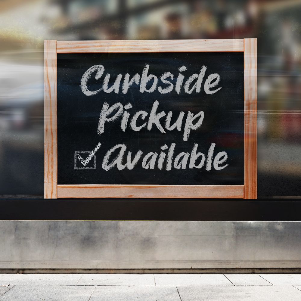 Chalkboard sign in a shop window reads “Curbside Pickup Available” with a checkmark