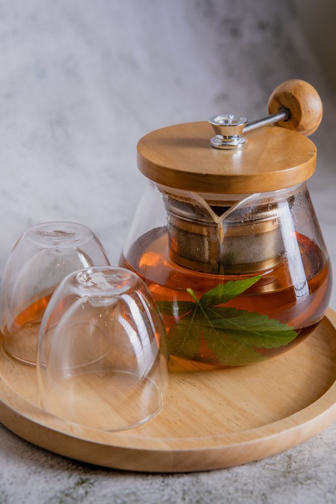Glass teapot with orange tea and mint leaves, with two upside-down glasses on a wooden tray.