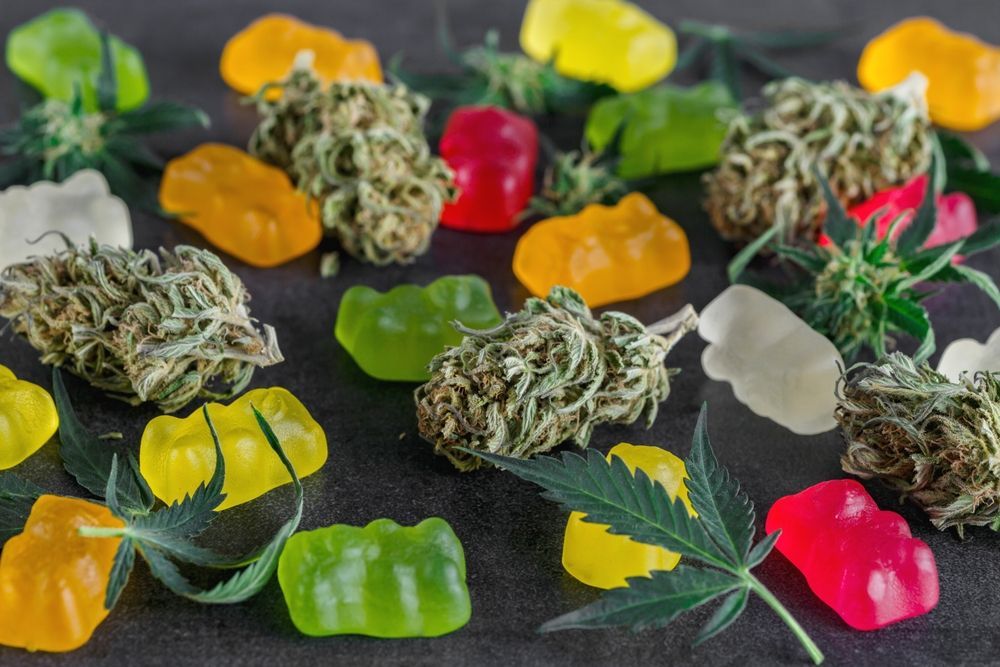 Colorful gummy candies and cannabis buds arranged on a dark surface with cannabis leaves.