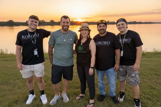 Five friends smiling outdoors at sunset near a lake, standing on grass in casual clothes.