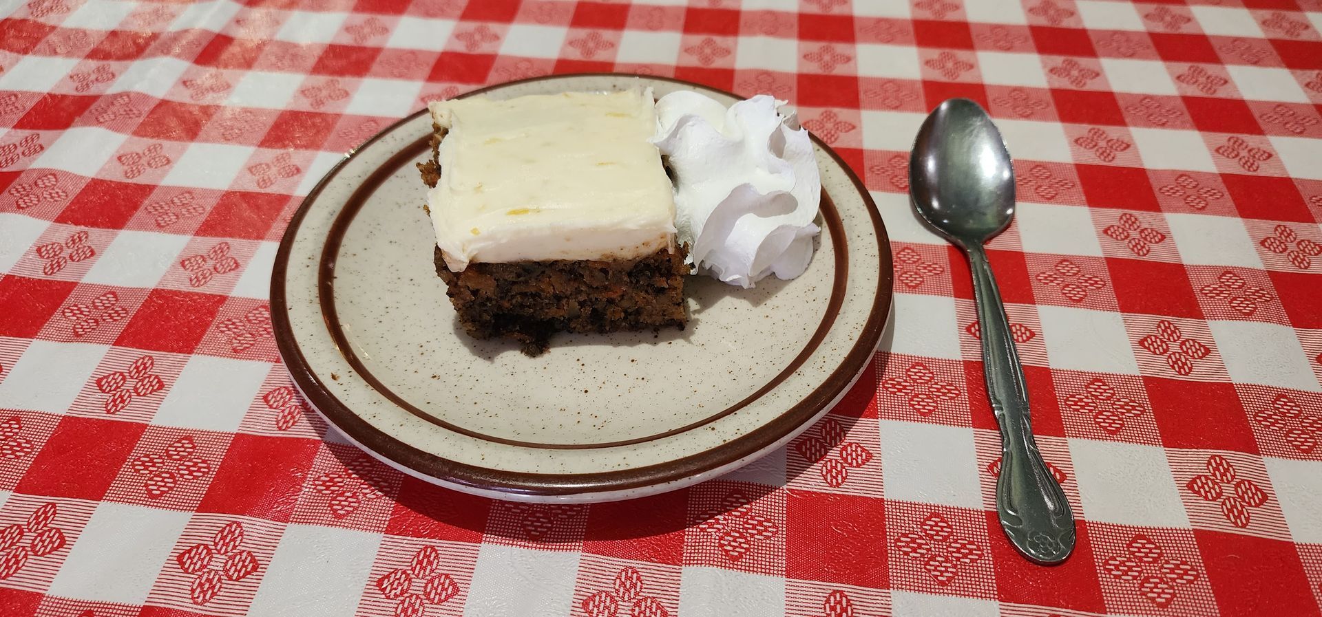 Slice of chocolate cake with white frosting on a plate, with whipped cream and a spoon on a red checkered tablecloth