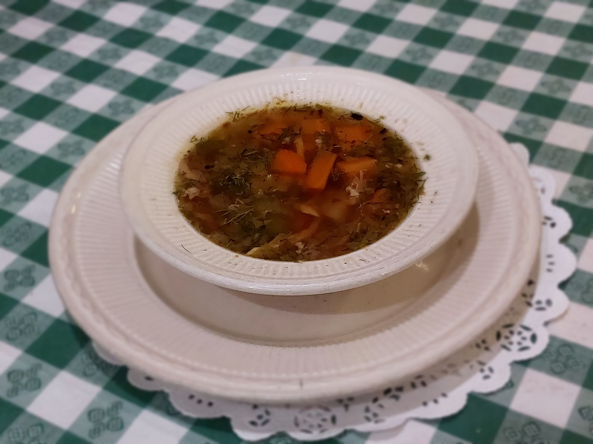 Bowl of vegetable soup on a white plate over a green checkered tablecloth