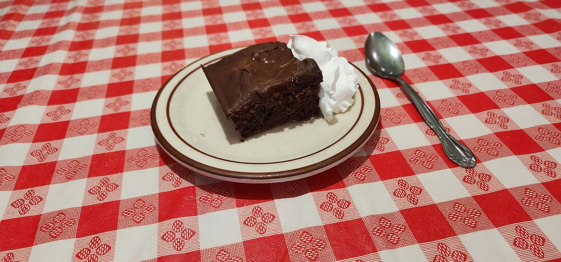 Chocolate cake slice on a plate with whipped cream and a spoon on a red checkered tablecloth