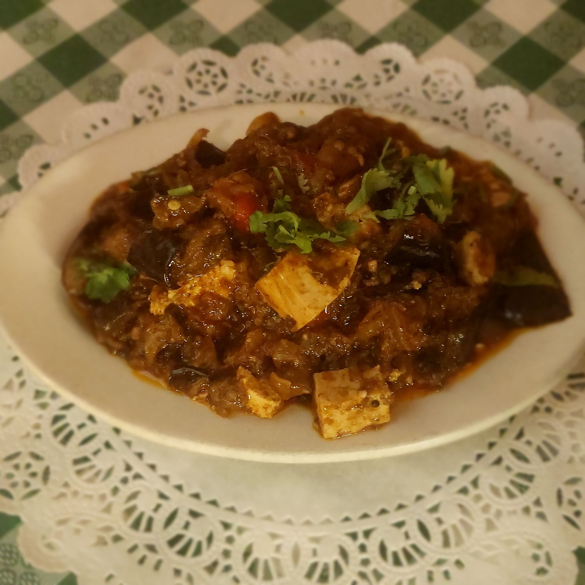 Plate of dark curry topped with herbs, served on a white doily-lined plate on a checkered tablecloth