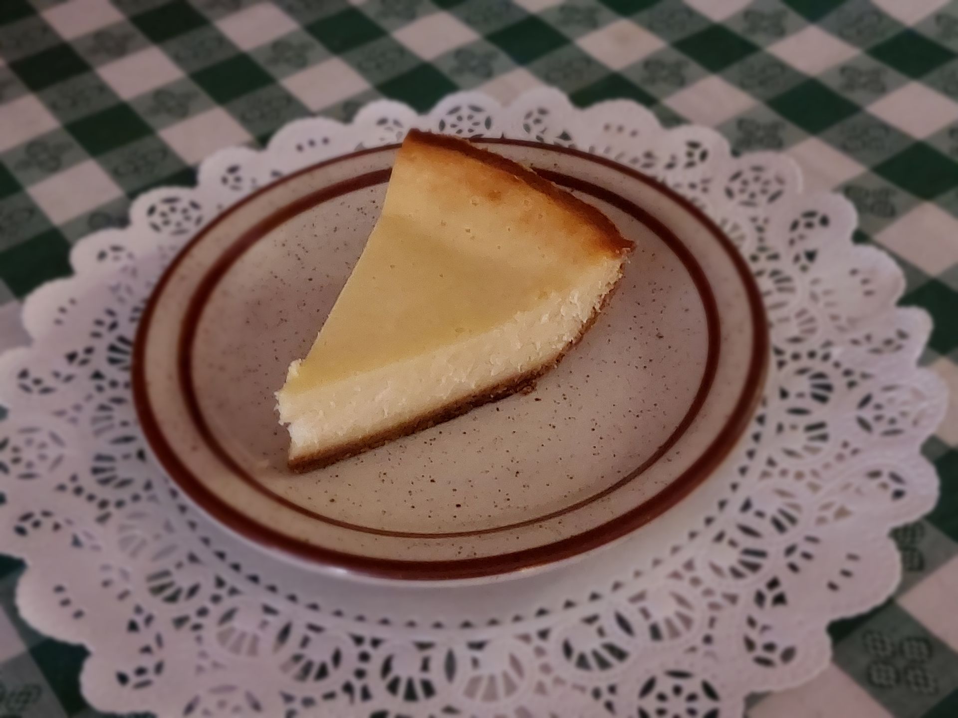 Slice of cheesecake on a brown-rimmed plate atop a lace doily on a green checkered tablecloth