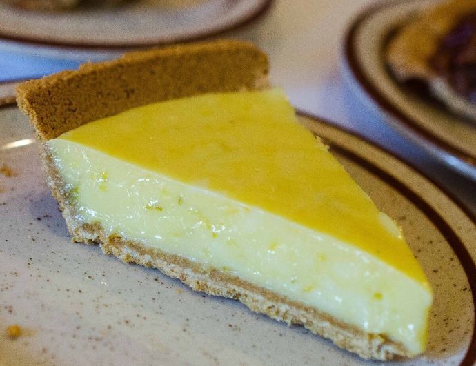 Slice of yellow-topped cheesecake on a plate, with a graham cracker crust and blurred pastries in the background