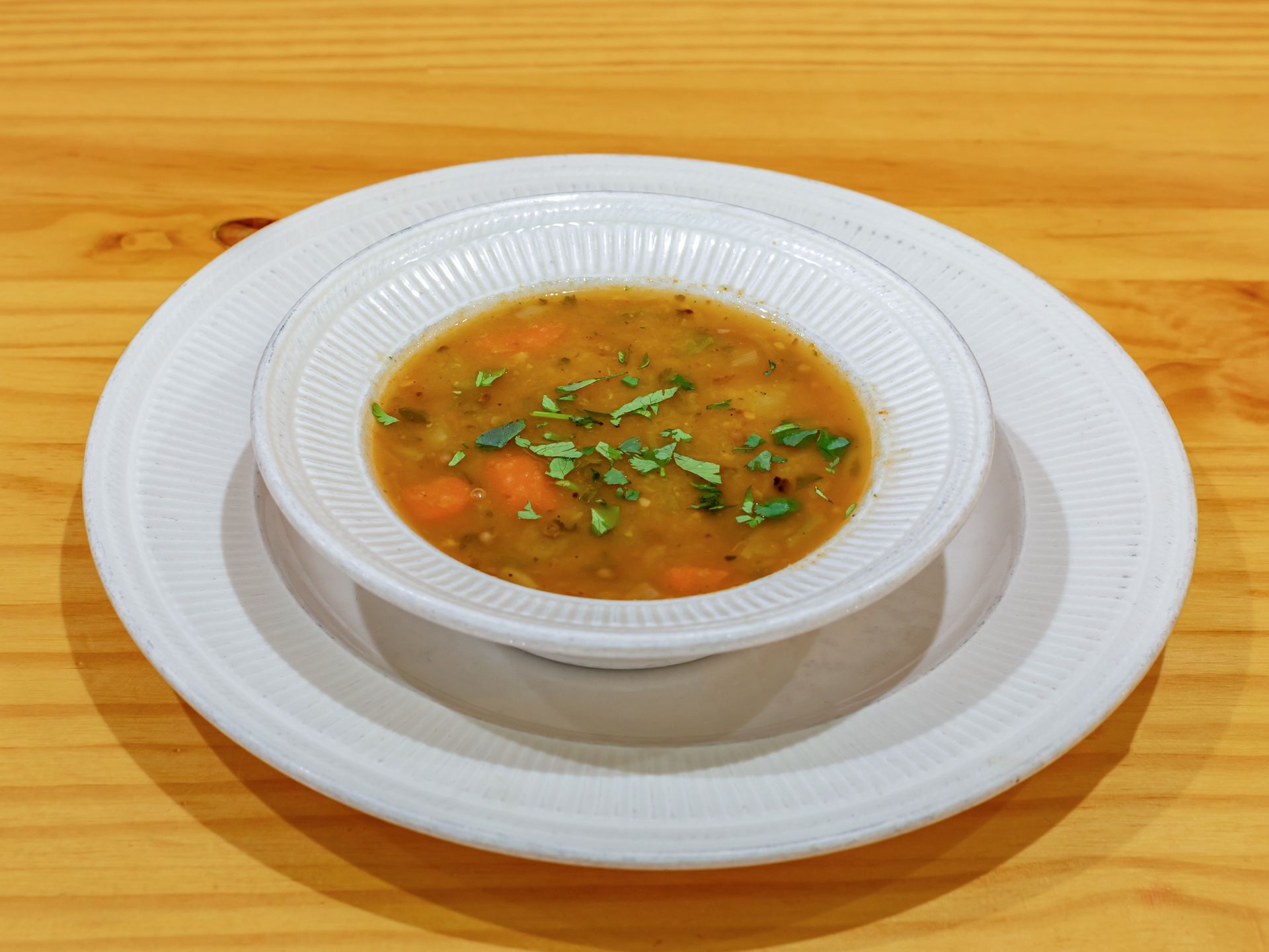 A bowl of vegetable soup garnished with herbs on a white plate, set on a wooden table.