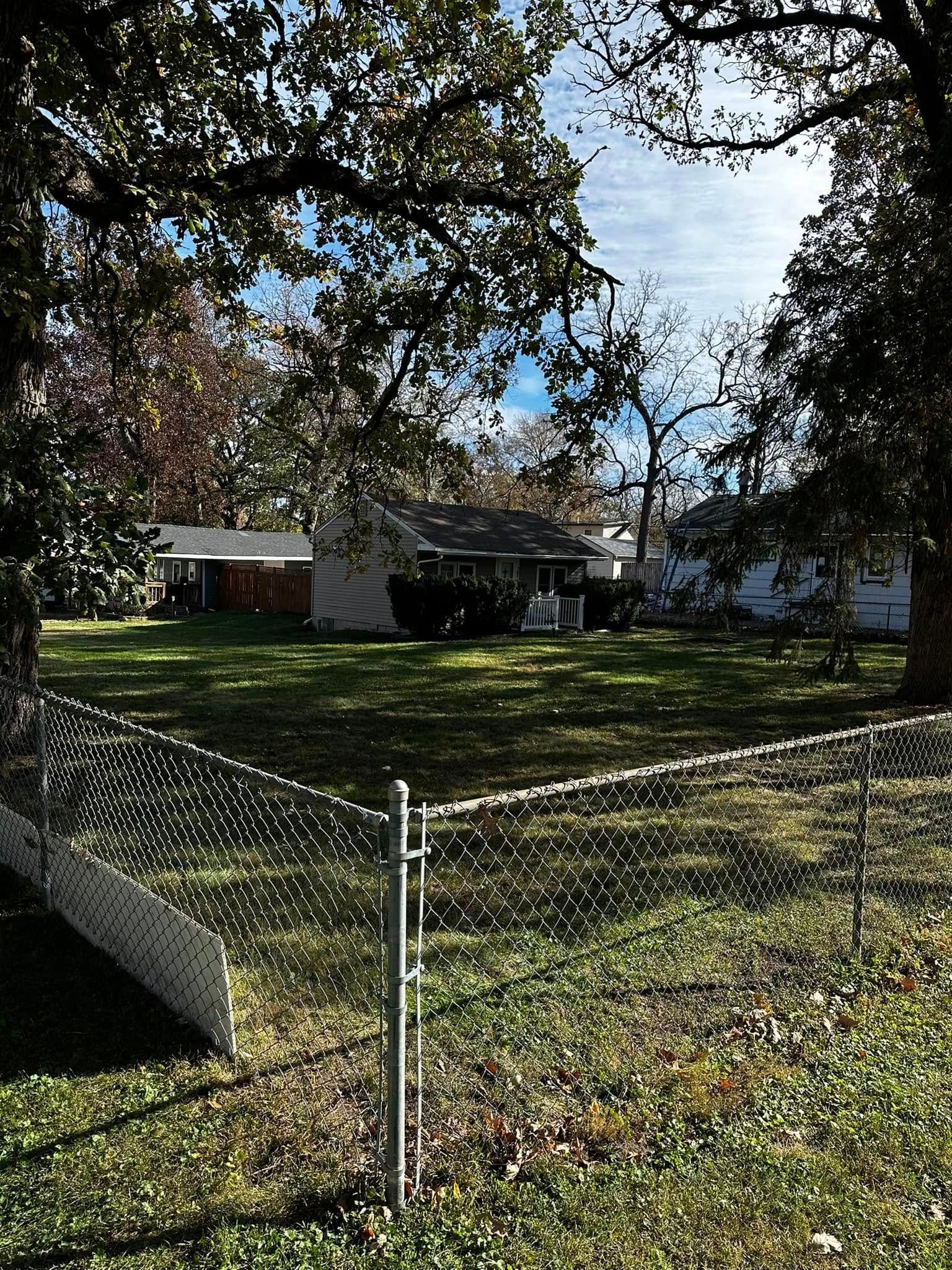 Chain-link fence in a grassy yard under large trees, with houses in the background.