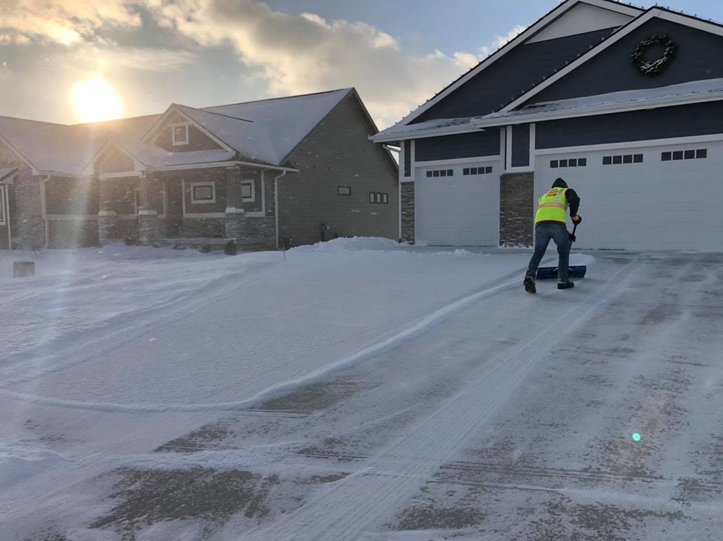 Snow-covered suburban driveway with a worker in a high-visibility jacket shoveling near a garage at sunrise