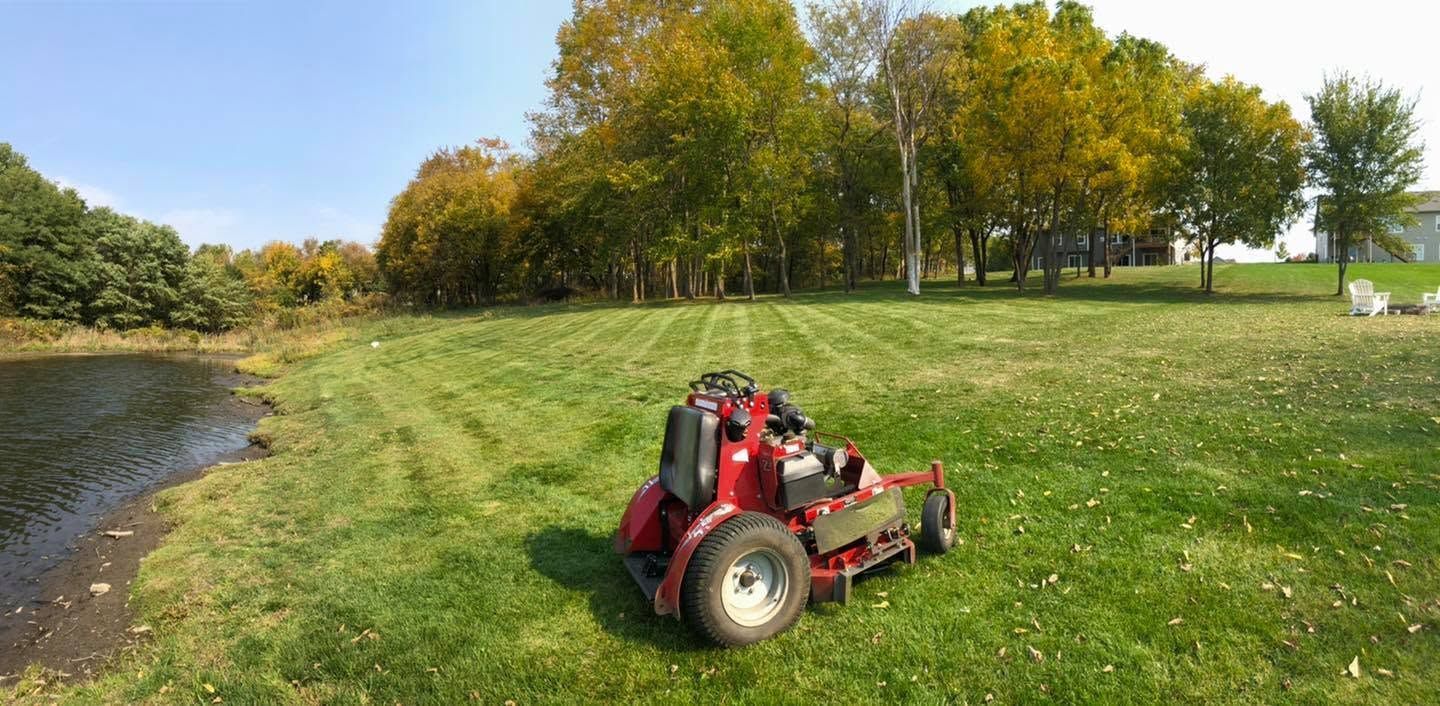 Red mower on a grassy field by a pond with autumn trees in the background