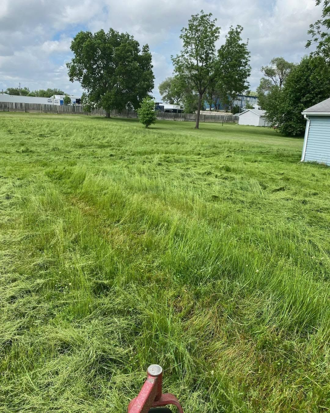 Wide grassy yard with trees and a white shed under a cloudy sky; a red sprinkler in the foreground