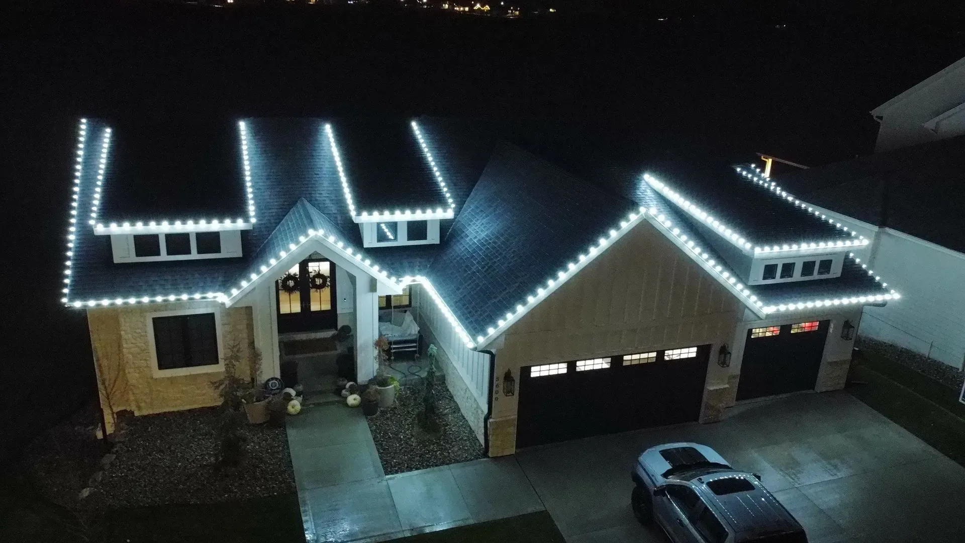 Night view of a house and garage outlined with white Christmas lights, with a car parked in the driveway.