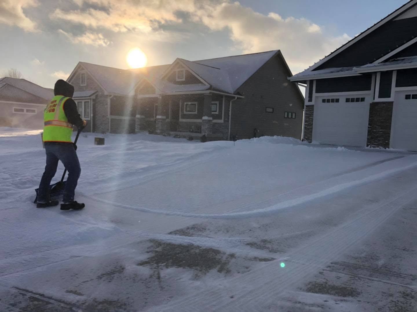 Worker in reflective vest stands on a snowy driveway beside suburban houses at sunset.