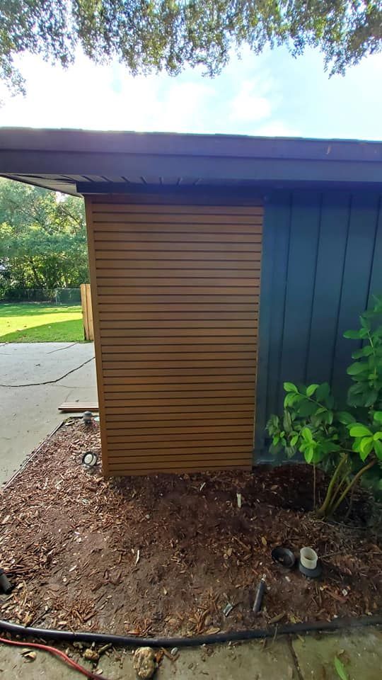 A modern house exterior featuring a section of horizontal wooden slats adjacent to dark vertical siding by a mulch bed.