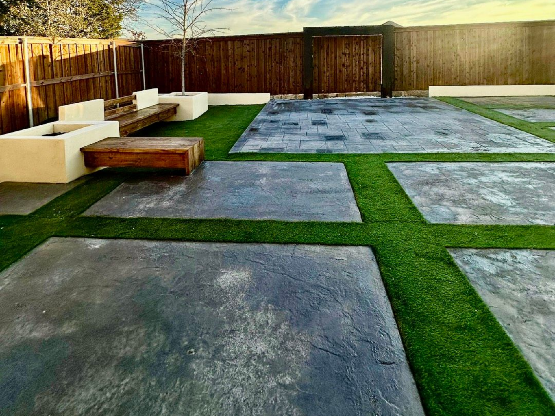 Backyard patio with square stone pavers and artificial grass, wooden bench, and wooden fence under a blue sky. Backyard patio with square stone pavers and artificial grass, wooden bench, and wooden fence under a blue sky.