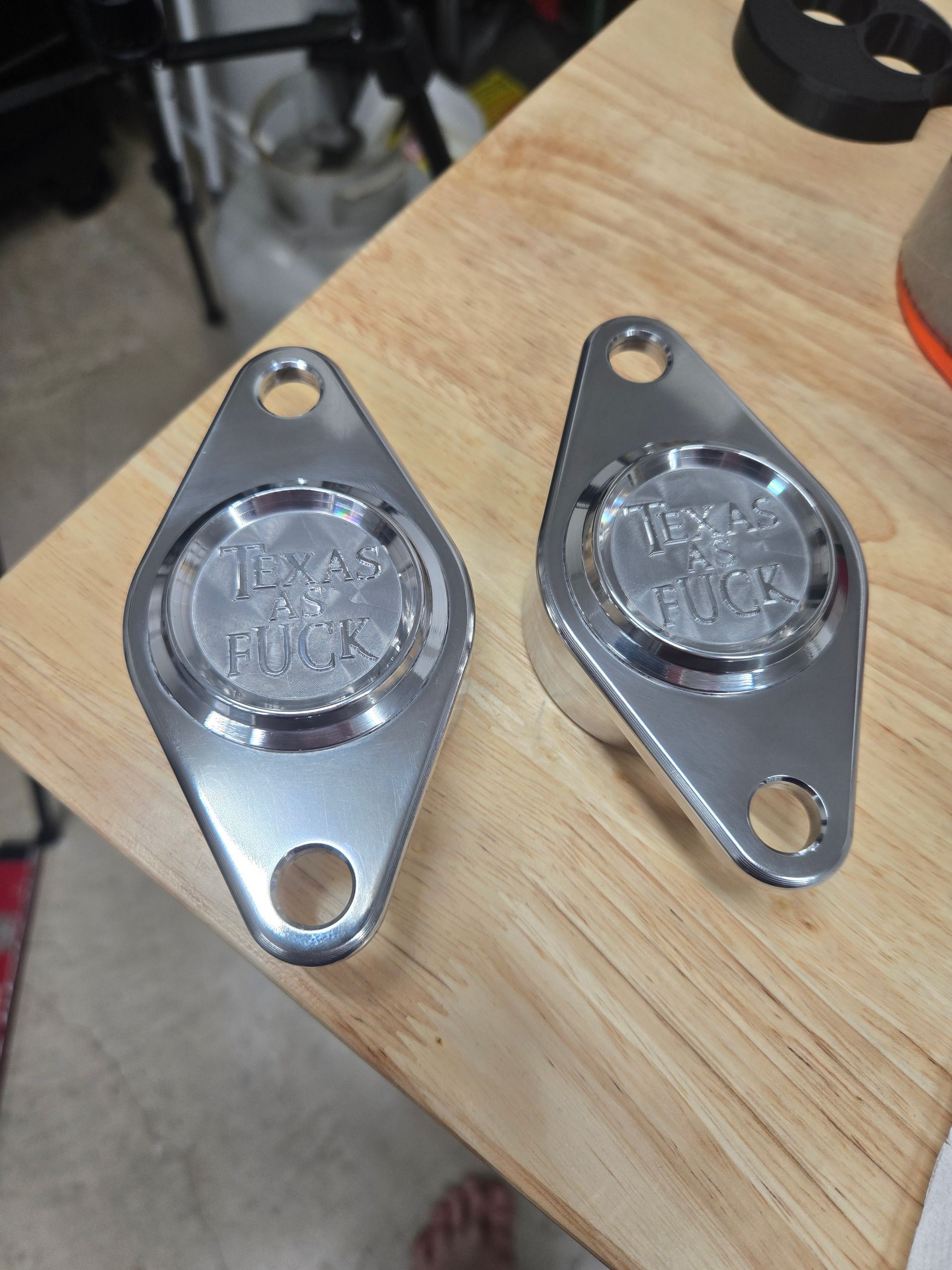Two polished metal motorcycle derby covers with visible text, sitting on a wooden surface.