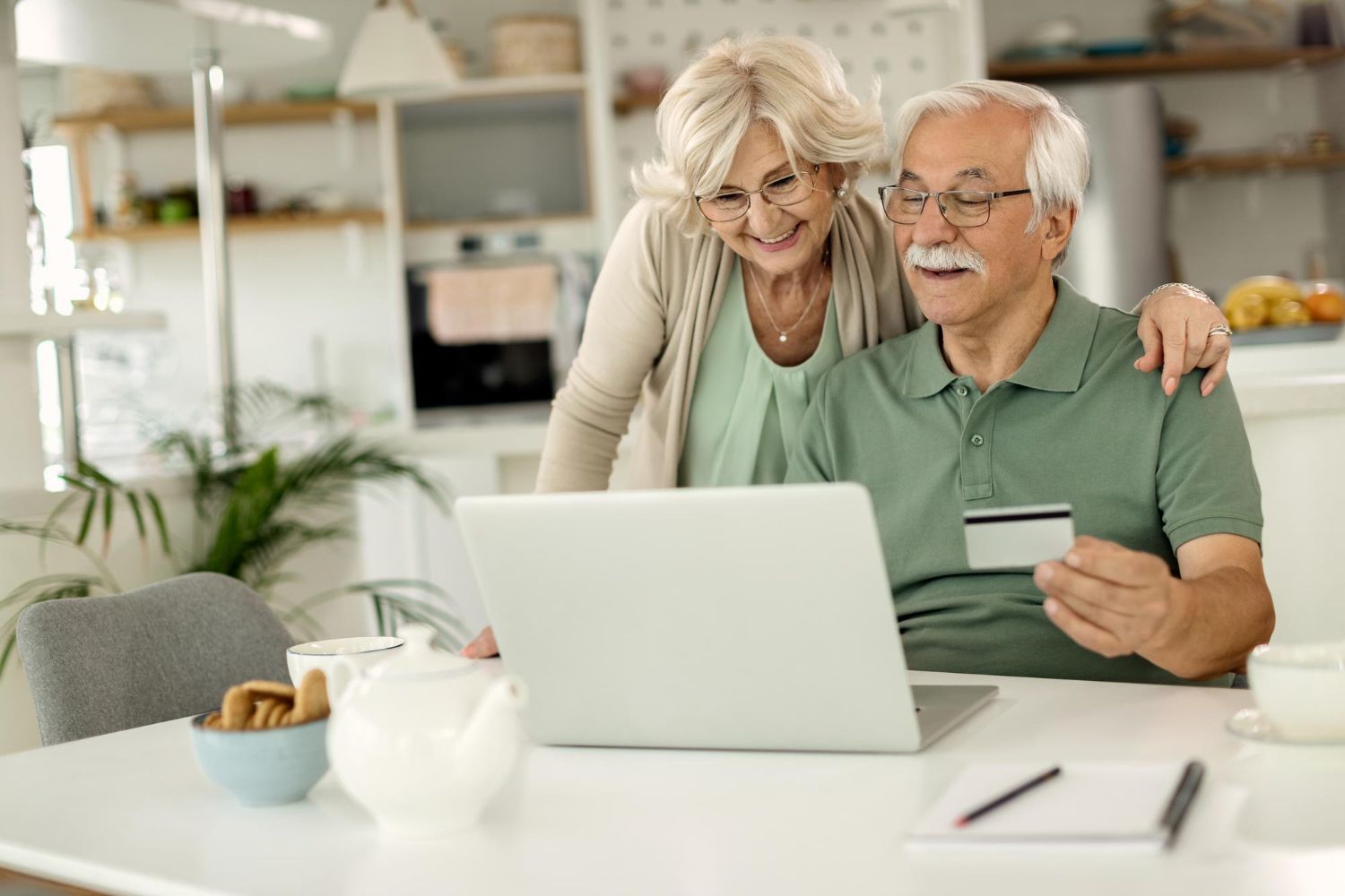 Older couple shopping online, man holding credit card, woman looking over his shoulder, laptop on table.