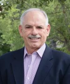 Man in suit and patterned shirt smiles in front of green foliage.