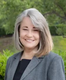 Woman in gray blazer smiles outdoors with blurred greenery.