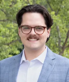 Man in a navy blazer and white shirt smiles outdoors near green foliage.