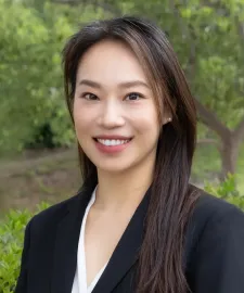 Woman in black blazer smiling at the camera, trees in background.