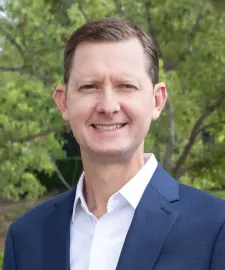 Man in a navy blazer and white shirt smiles outdoors near green foliage.