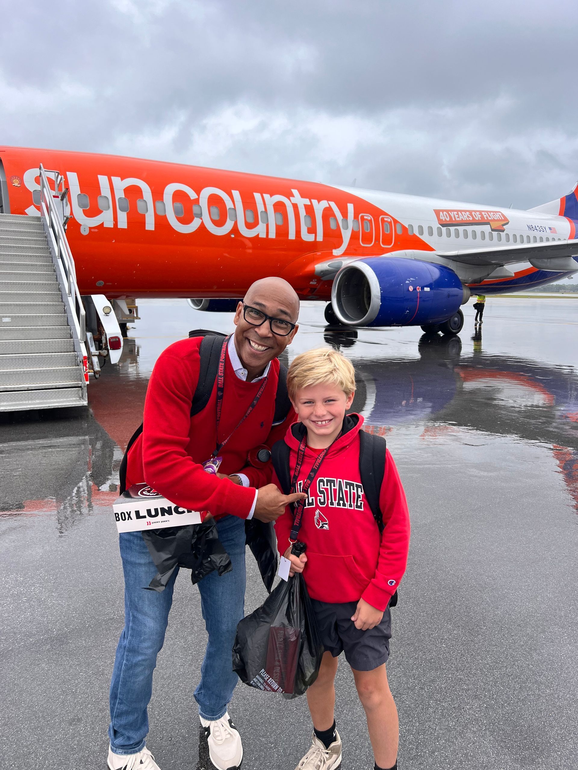 Man points at child wearing red hoodie in front of a Sun Country airplane; gray sky.