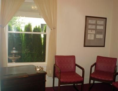 Small office waiting area with two red chairs, framed notice, and window with curtains overlooking greenery