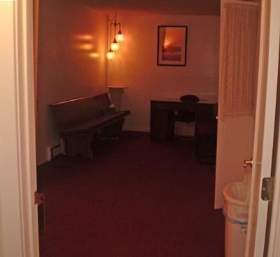 Dimly lit room with a piano, desk, wall lamp, and open doorway on a red carpet.