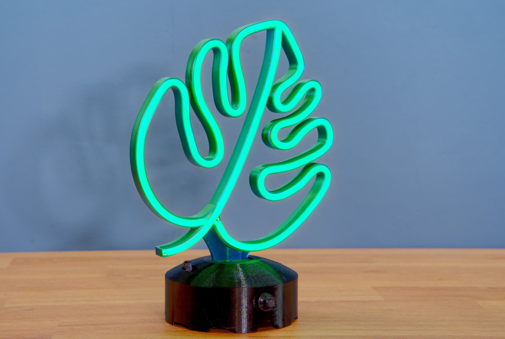 A glowing, bright green LED monstera leaf sign stands on a circular, dark gray textured base against a blue wall.