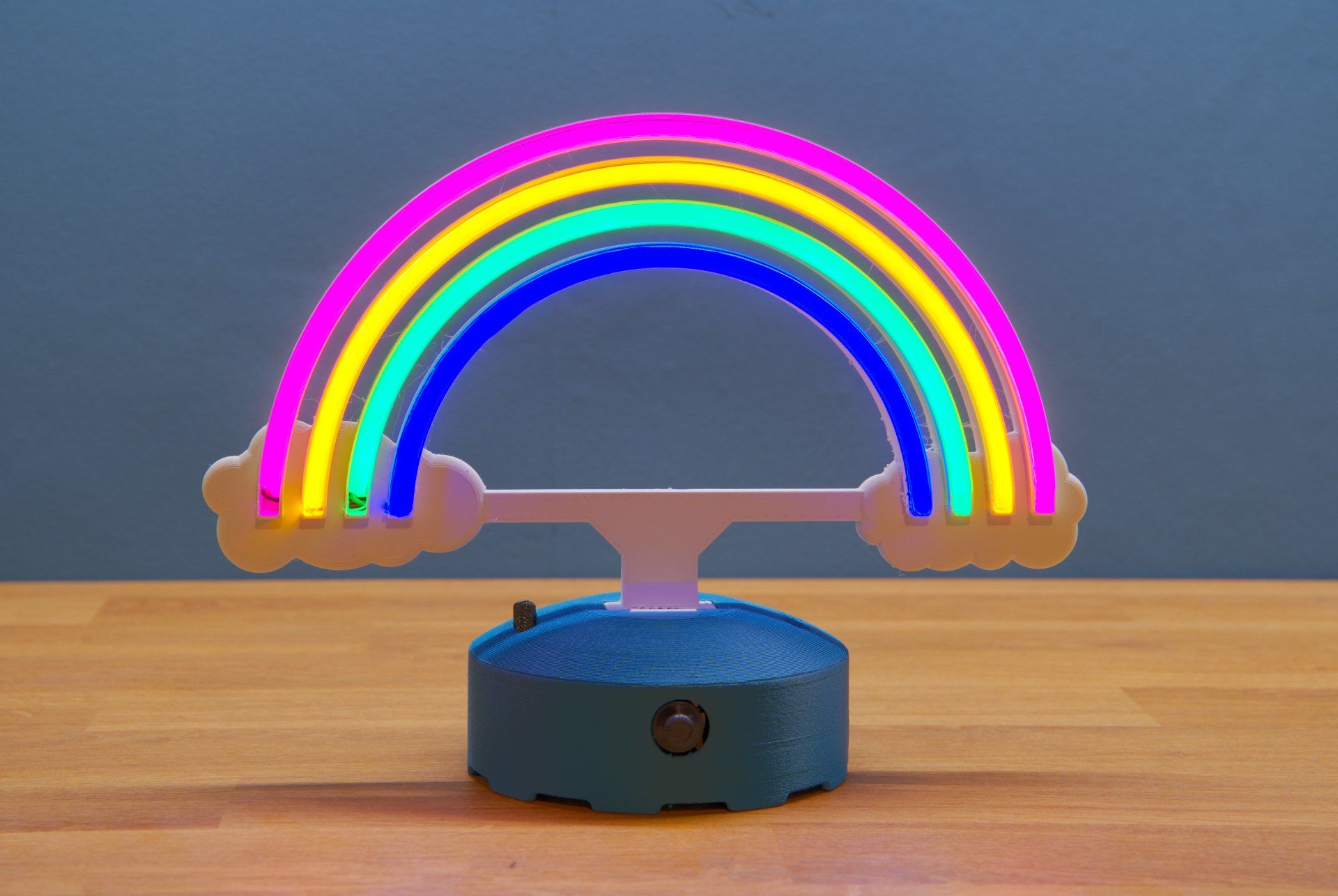A glowing, multi-colored LED rainbow arch table lamp with two cloud-shaped ends, mounted on a blue base on a wood surface.