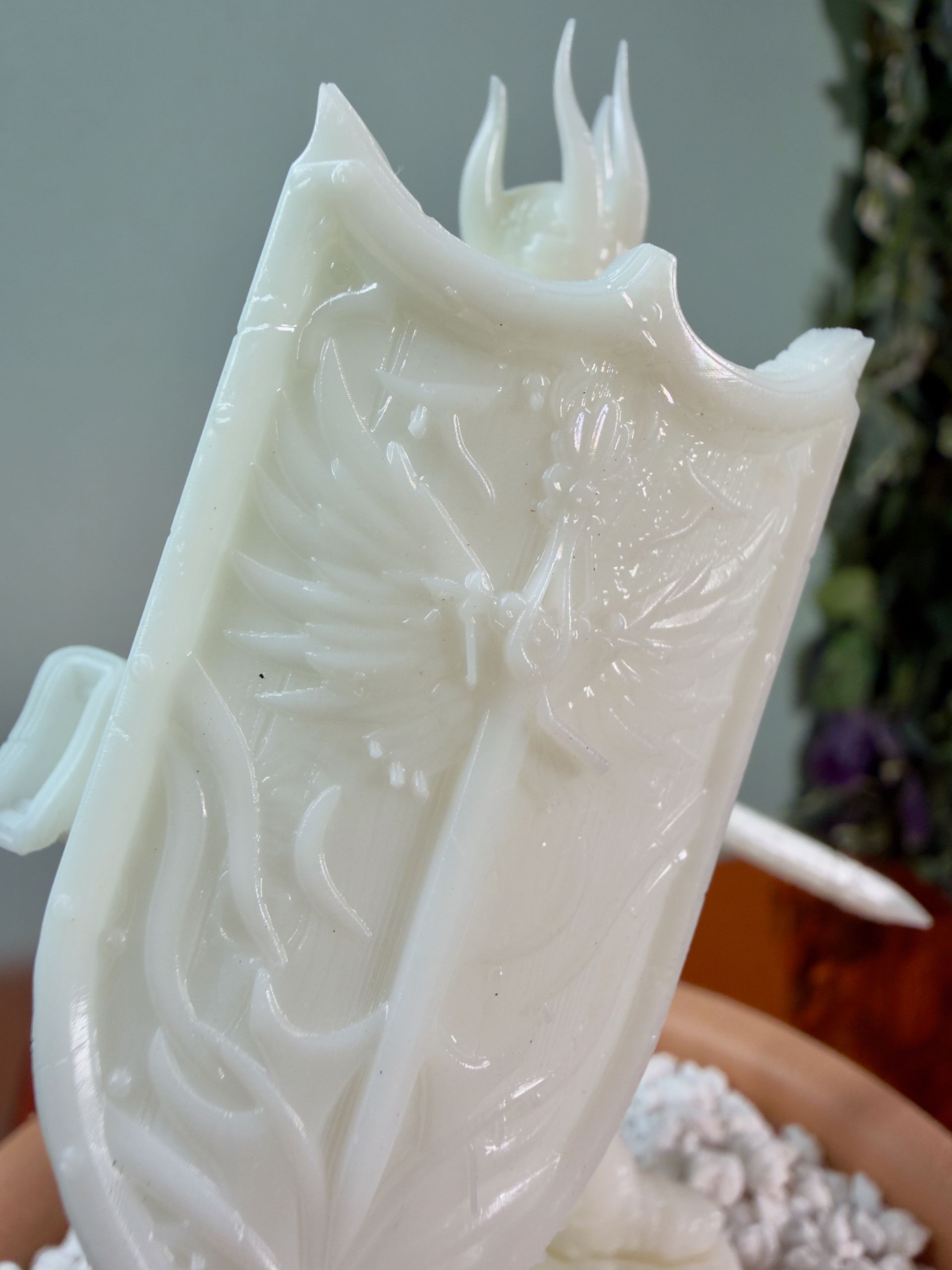A white, 3D-printed shield featuring a phoenix emblem and flame details, held in a blurry, natural indoor setting.