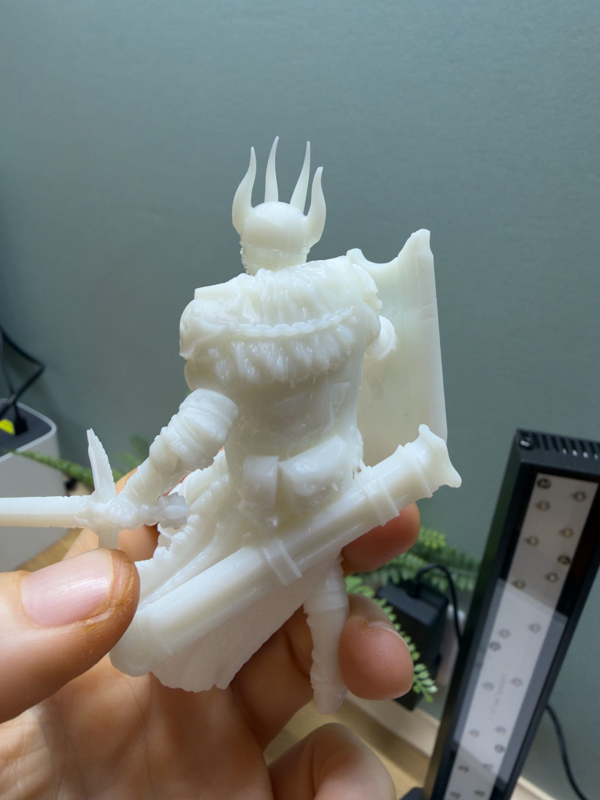 A hand holding a white 3D-printed miniature figure of a fantasy knight in armor with a sword and shield.