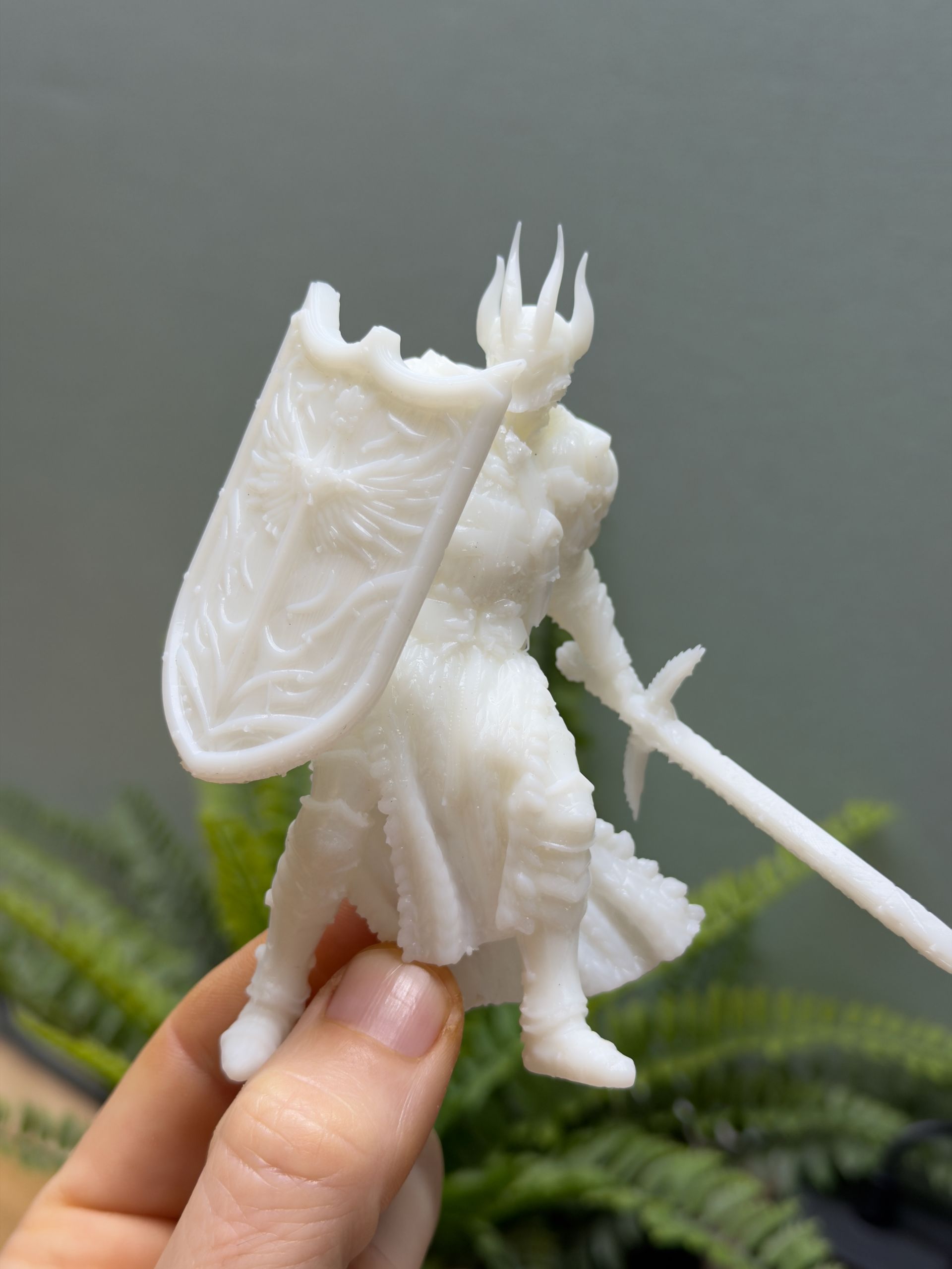 A hand holds a white, 3D-printed miniature figure of a fantasy knight wearing a spiked helmet, holding a shield and sword.