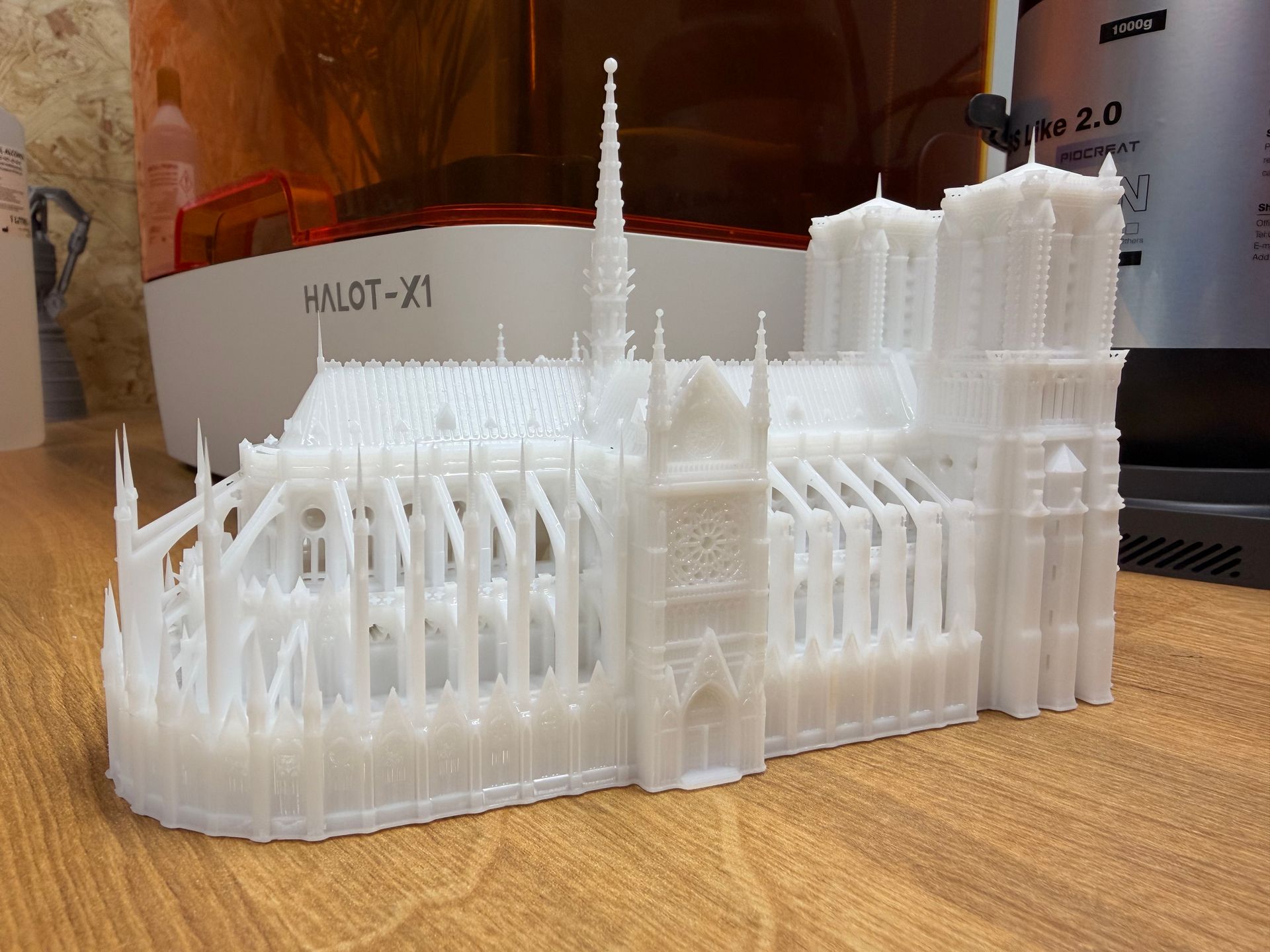 A detailed, white 3D-printed model of Notre-Dame Cathedral sits on a wooden desk next to a resin printer.
