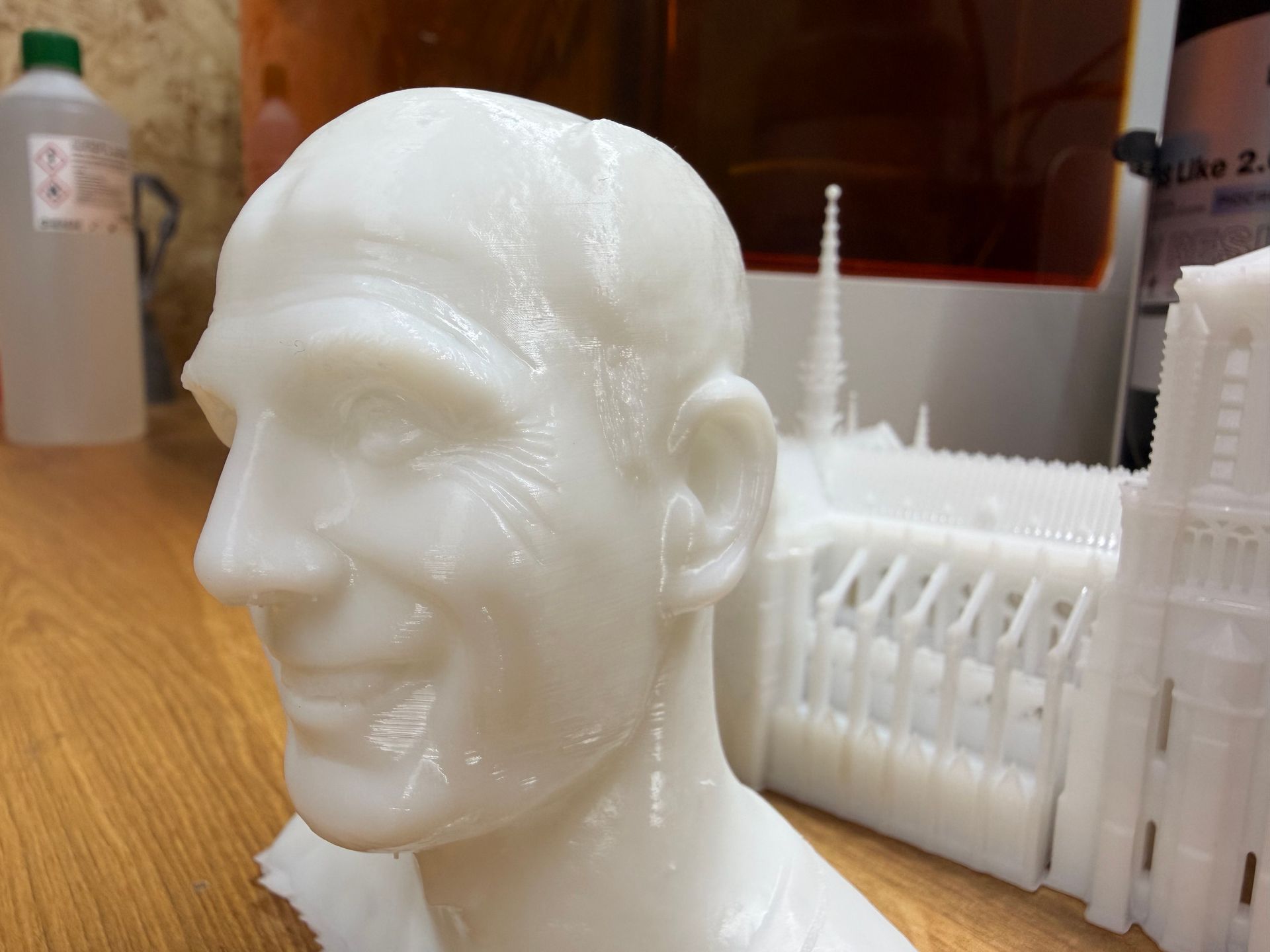 A 3D-printed white sculpture of a head next to a 3D-printed model of a cathedral on a wooden surface.