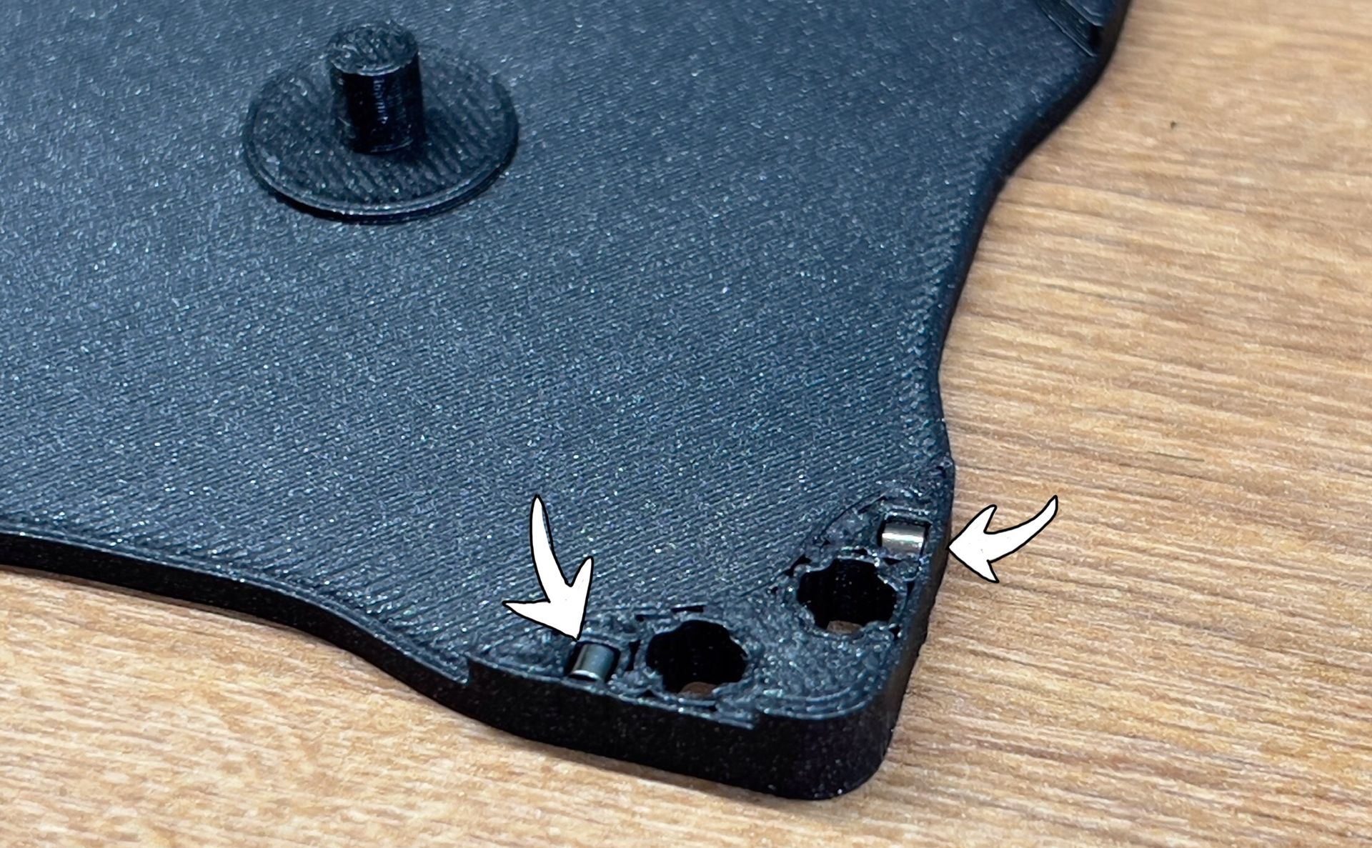 A black 3D-printed part sits on a wooden surface, with two white arrows pointing to small, clipped plastic support nubs.