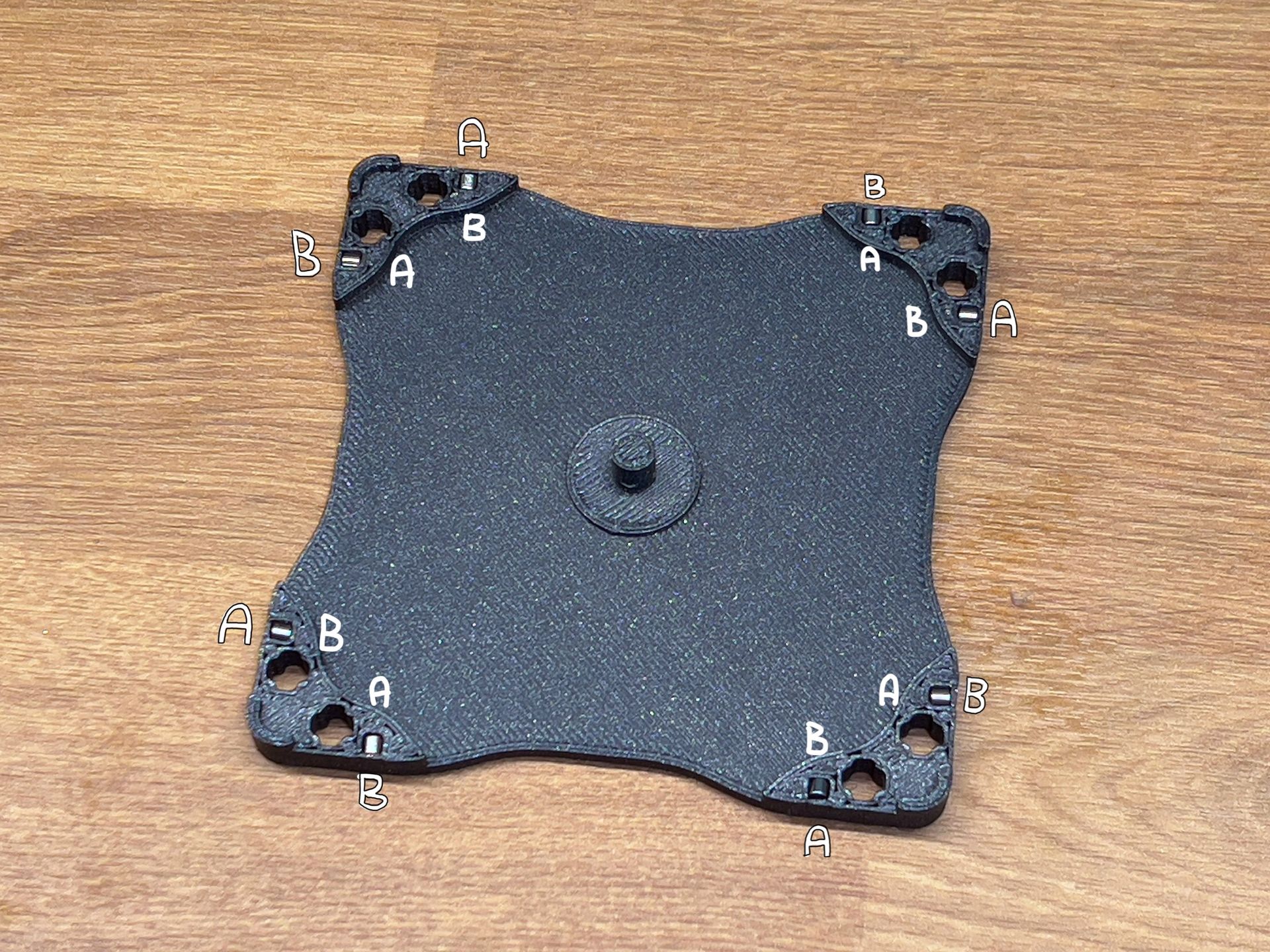 A black, square 3D-printed mounting plate with corner holes labeled A and B, resting on a wooden surface.